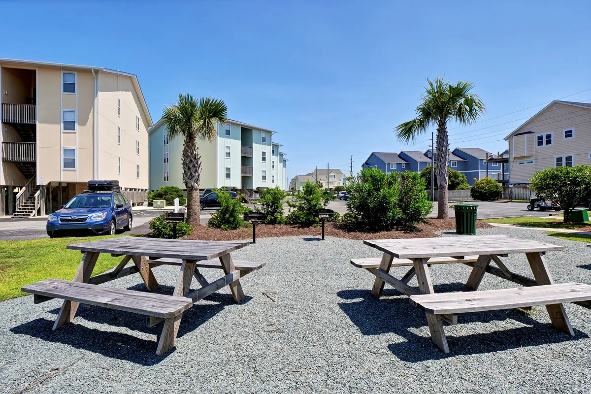 An inviting exterior scene featuring picnic tables shaded by palm trees, surrounded by well-kept greenery. The area is ideal for grilling, al fresco dining, and casual gatherings with friends or neighbors.