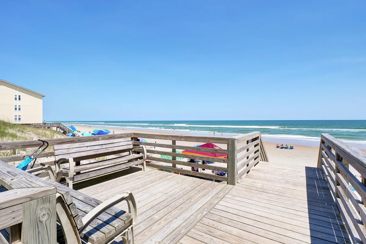 A spacious beach deck with chairs and umbrellas, offering a panoramic view of the ocean and sandy shore below. The setup hints at leisurely sunbathing, beachcombing, or simply soaking up the coastal atmosphere together.