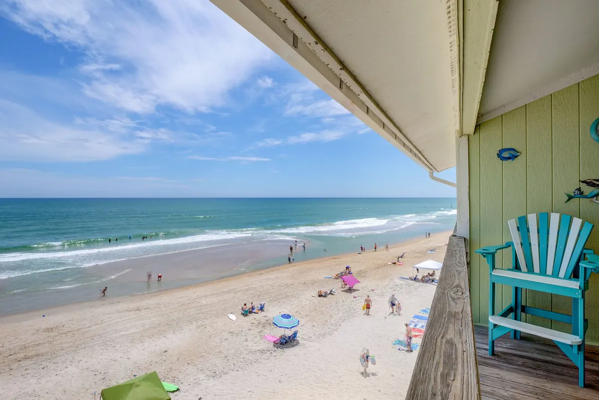 The image captures a sweeping beachfront scene as viewed from the condo's balcony. Gentle waves roll onto the golden sand, dotted with colorful beach umbrellas and a turquoise chair, evoking relaxation and fun. The inviting setting hints at days filled with sunbathing, swimming, and vibrant beach activity, all just steps from your door.