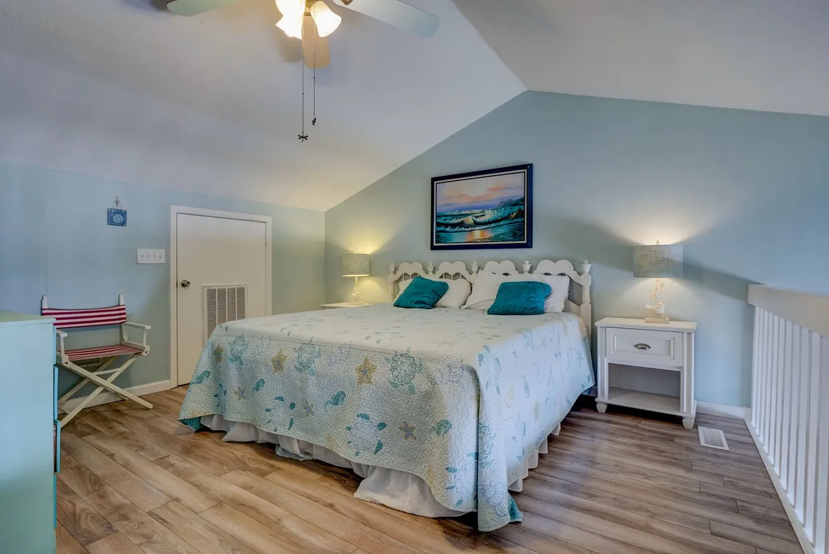 A serene bedroom featuring a bed with a decorative coverlet, framed artwork, nightstands with lamps, and a comfortable seating area. Sunlight streams in, creating a calm and inviting atmosphere ideal for relaxation or reading.