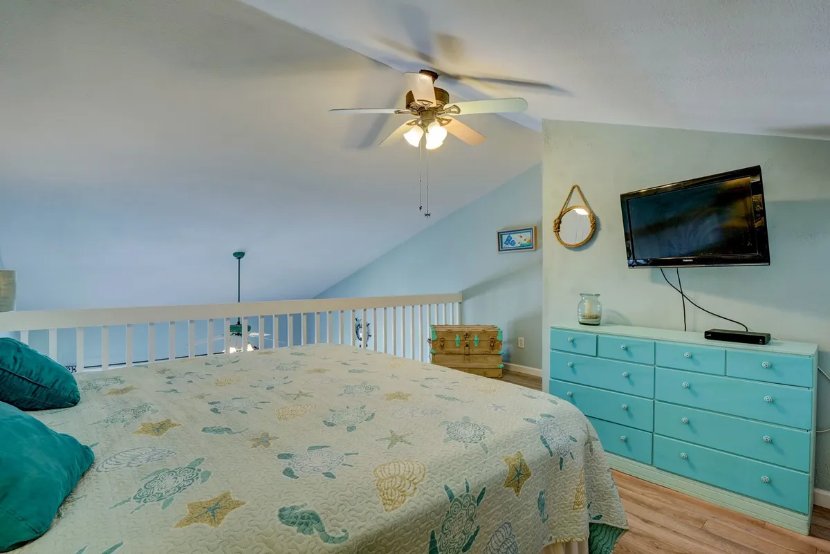 A serene, beachy bedroom featuring a king-size bed with a sea-themed quilt, turquoise dresser, wall-mounted television, and ceiling fan. Natural light brightens the space, creating a cheerful yet restful vibe ideal for a flexible sleeping area.