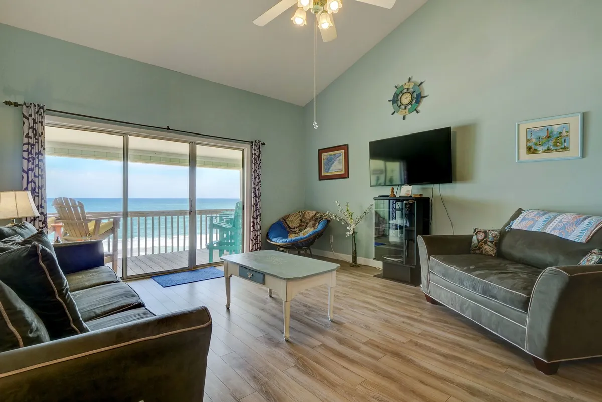 A bright, open living room with a sofa set facing large sliding glass doors that lead out to a balcony. The coffee table and decorative wall art add visual interest, while sunlight floods the space, drawing attention to the expansive ocean view visible through the glass.