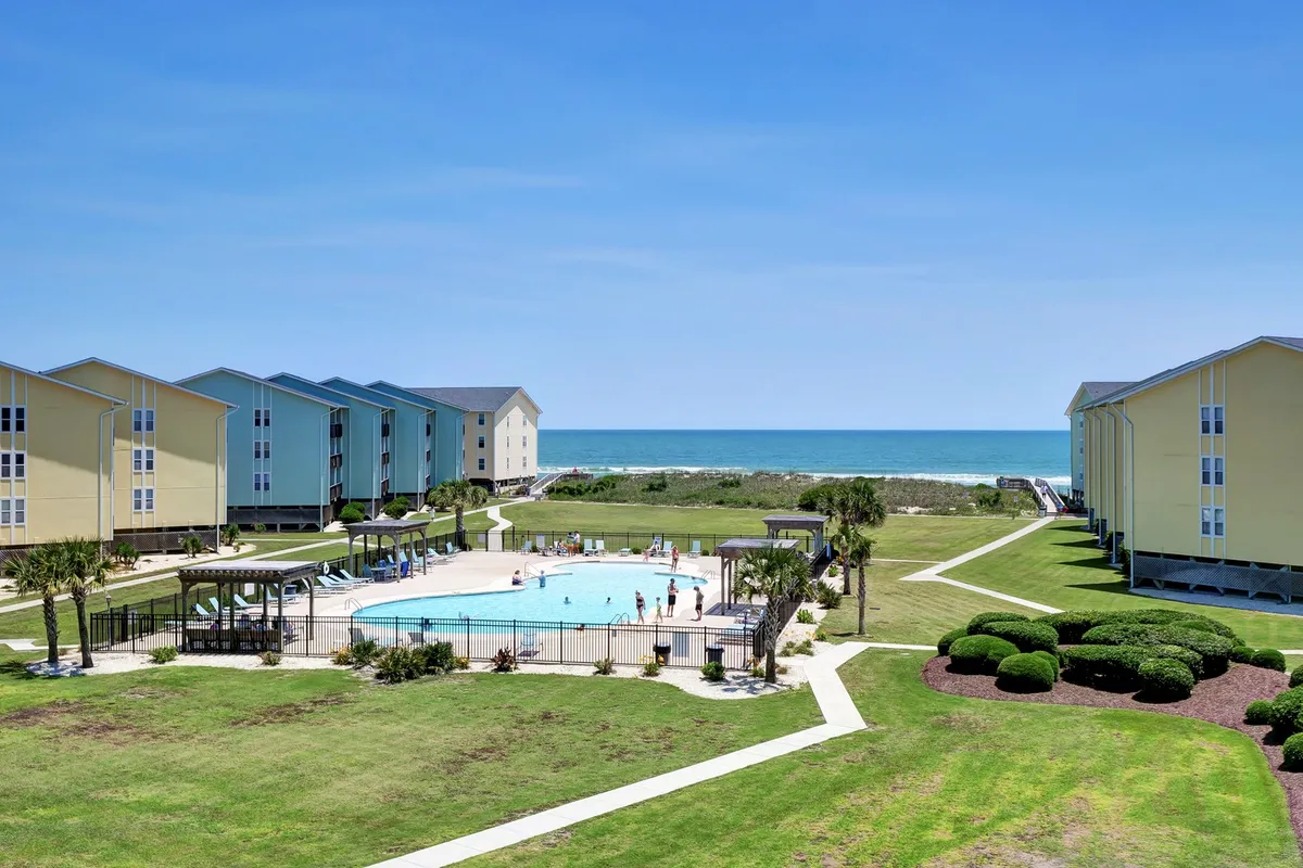 An outdoor swimming pool with clear blue waters sits overlooking the ocean, surrounded by landscaped greenery and resort buildings. The setting invites relaxation, swimming, and sunbathing, offering families an alternative to the beach.