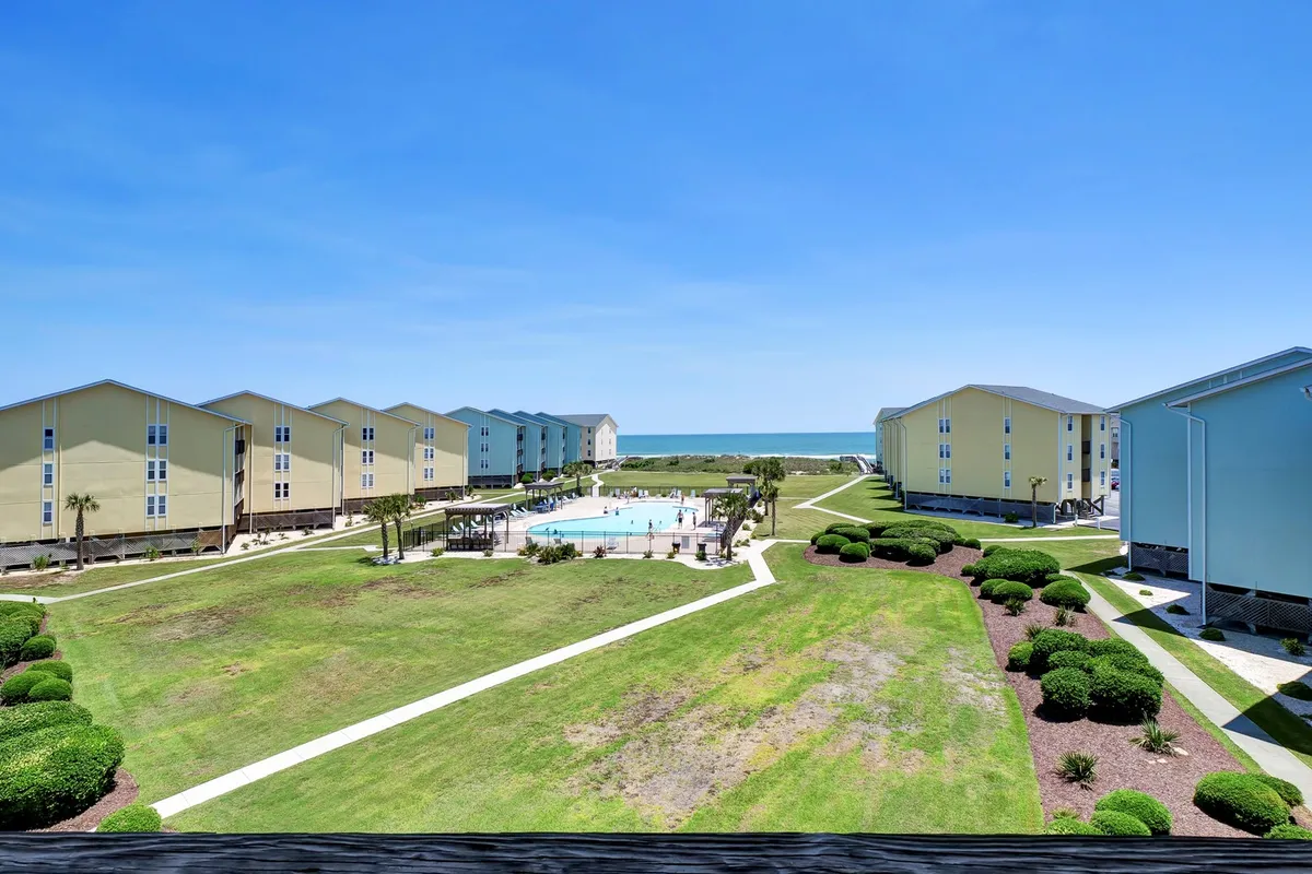 A vibrant exterior shot capturing the community swimming pool set against landscaped lawns with an ocean view and residential buildings in the background. Lounge chairs are arranged poolside, inviting relaxation and social interaction in a sun-drenched setting.