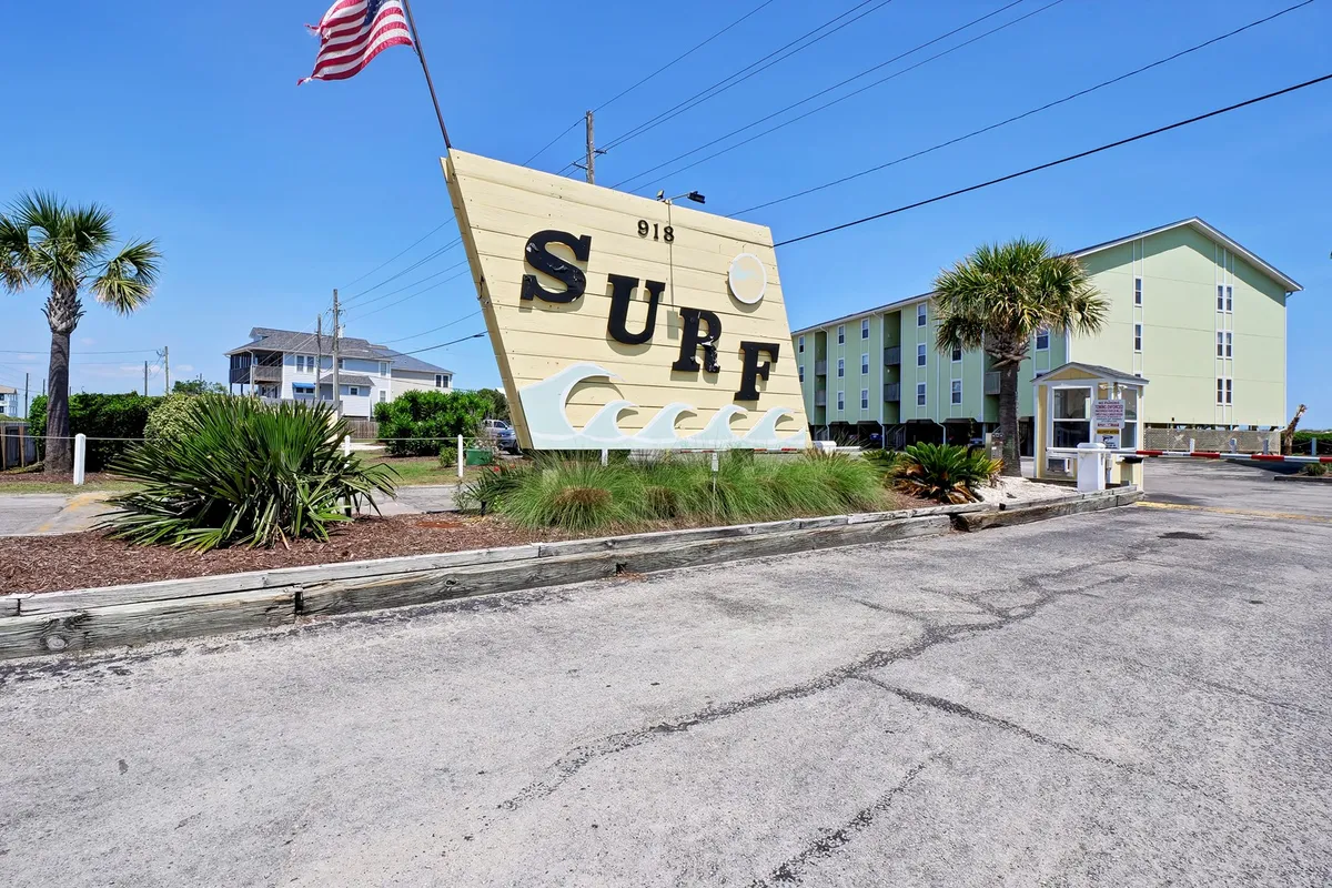 This exterior image features a prominent 'SURF' sign set among palm trees, with surrounding buildings visible, evoking a lively and playful beach community vibe. The scene suggests proximity to the ocean, communal outdoor areas, and a welcoming neighborhood atmosphere ideal for group vacations.