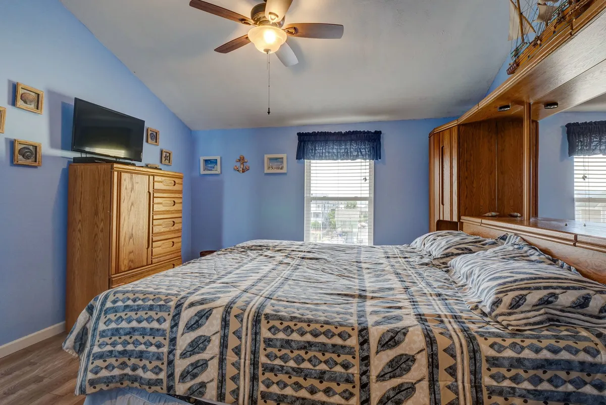 A tranquil, inviting king bedroom with a patterned bedspread, wooden dresser, wall-mounted TV, and decorative pictures above the bed. Soft daylight filters through a nearby window, highlighting the room's cozy atmosphere and ideal setup for restful nights or relaxing with a movie.