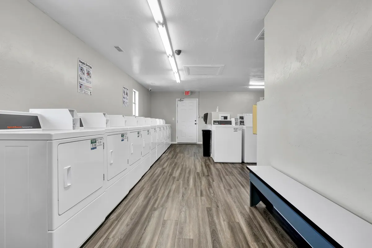 A clean, functional interior laundry area with rows of white washing machines, a bench for waiting, and a doorway to other building areas. The space is well-lit and efficient, emphasizing practicality and convenience.