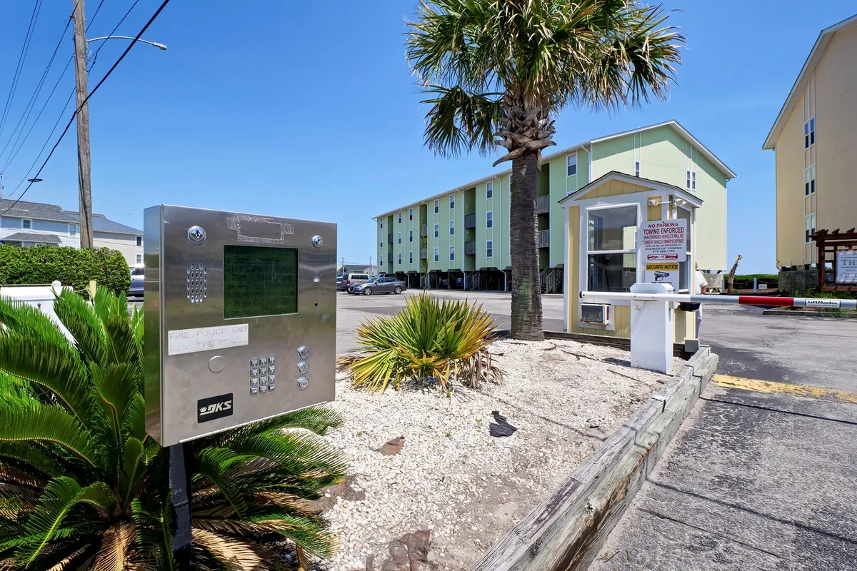 This image shows the building facade of Surf Condos, framed by palm trees and an entry gate, with an intercom system visible. The sunny exterior creates a relaxed, inviting mood and hints at the property's proximity to the beach and the communal atmosphere.