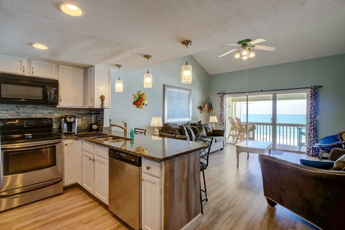 A bright interior scene showing a kitchen island with seating that looks out toward an ocean view, blending cooking and socializing spaces. The area is set up for group activities like preparing meals together and enjoying casual conversation with a scenic backdrop.