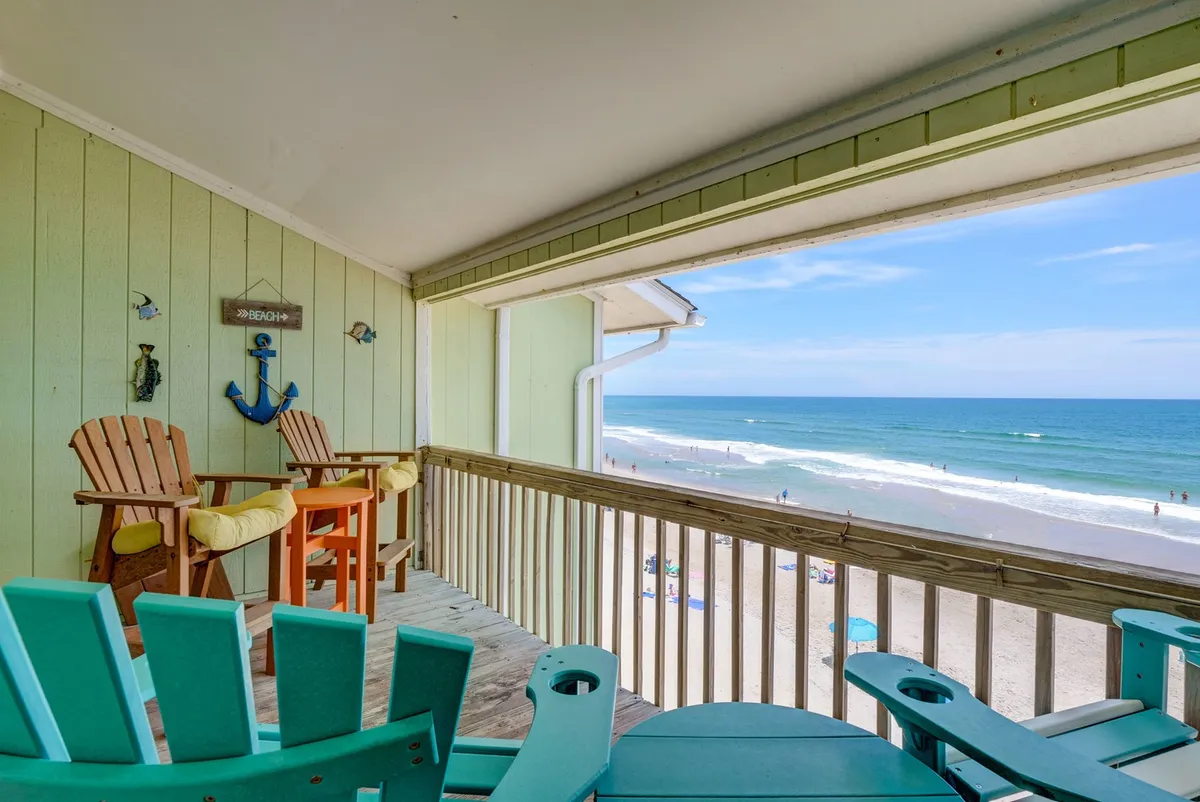 This exterior image features a serene beach view as seen from an oceanfront deck with Adirondack chairs and railing in the foreground. The tranquil atmosphere, soft early light, and the open expanse of the Atlantic Ocean visually support the idea of waking up to sunrises and enjoying intimate moments while overlooking the waves.