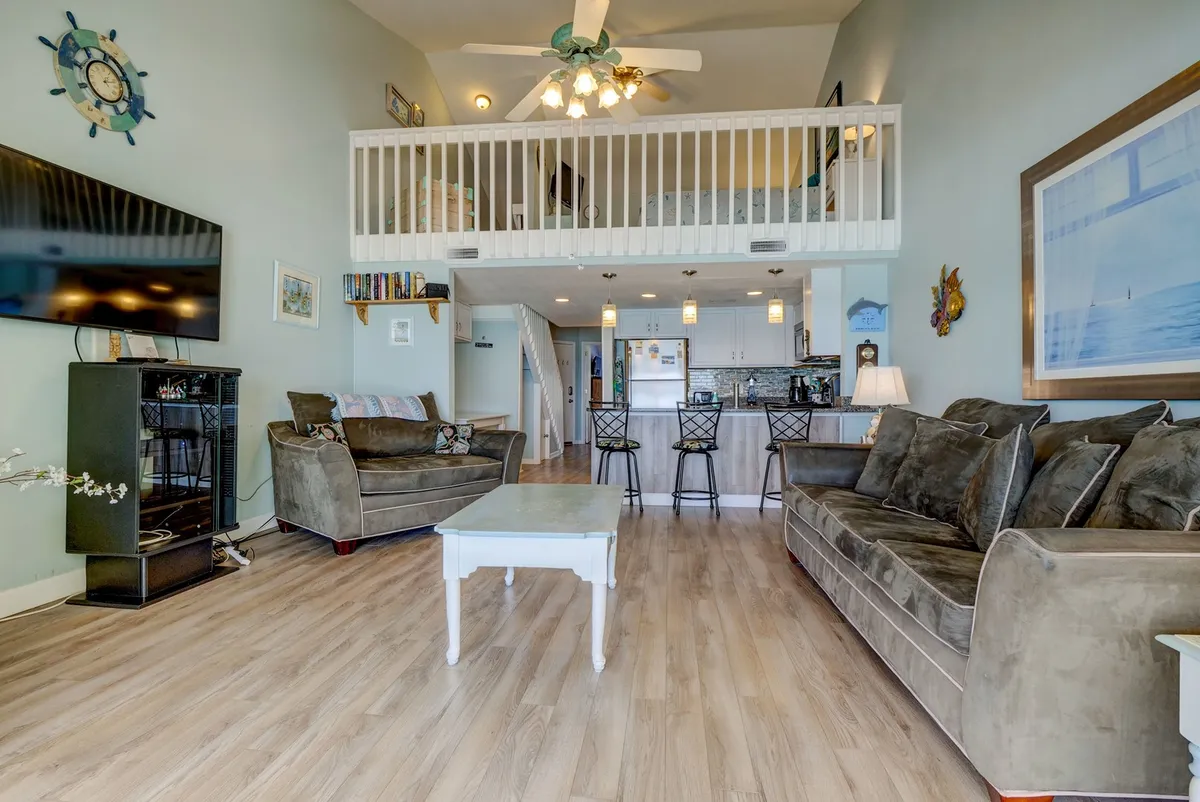 An inviting open-concept living area with comfortable sofas, a visible loft space, kitchen island, and large TV. The layout encourages both relaxing and socializing, with zones for movie nights, cooking, or hanging out as a group.