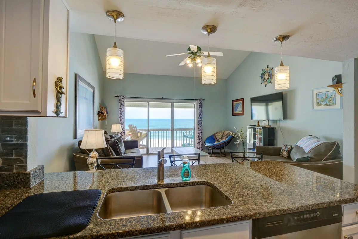 The image features a bright, modern living space with large windows offering sweeping ocean views, a kitchen island, and comfortable seating. The scene conveys a sense of relaxation and sociability, with natural light streaming in from the windows.